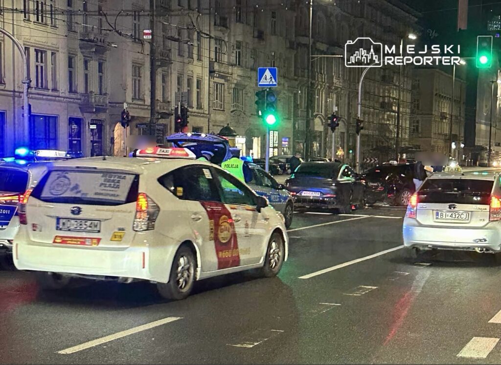 Radiowóz policyjny z włączonymi sygnałami świetlnymi blokuje ruch na ulicy, obok stoi taksówka.