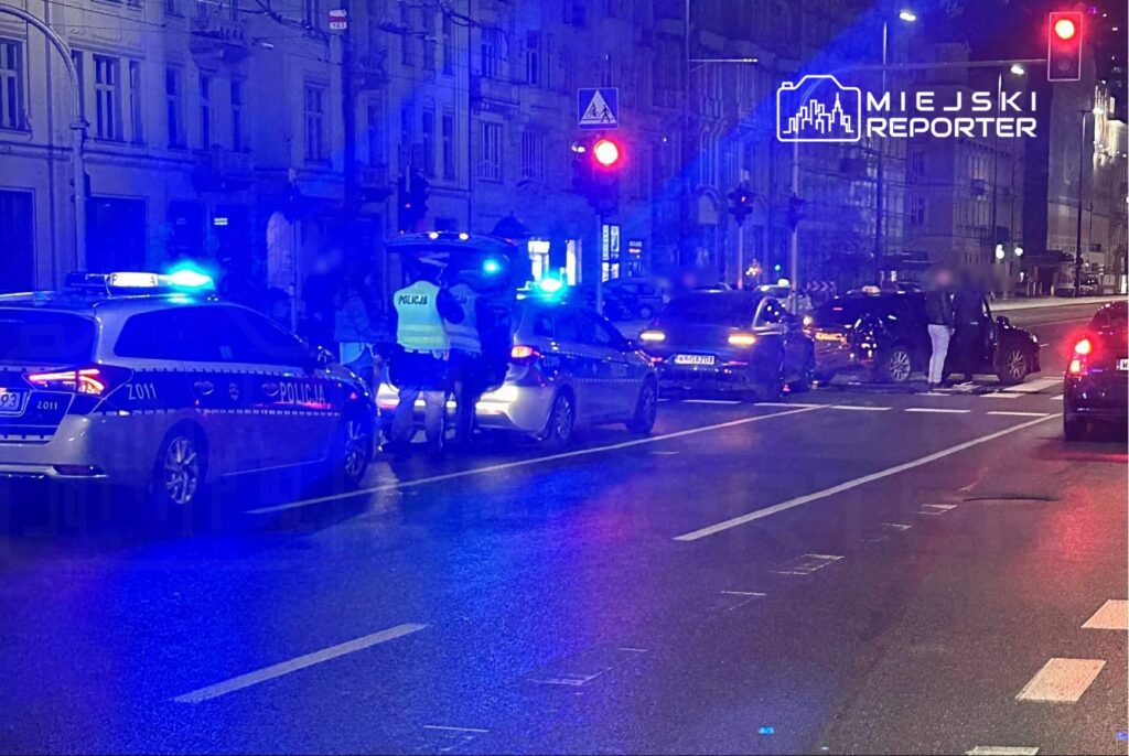 Na ulicy, w nocy, policyjne samochody z włączonymi sygnalizatorami zatrzymują pojazdy, a funkcjonariusze rozmawiają z kierowcami.