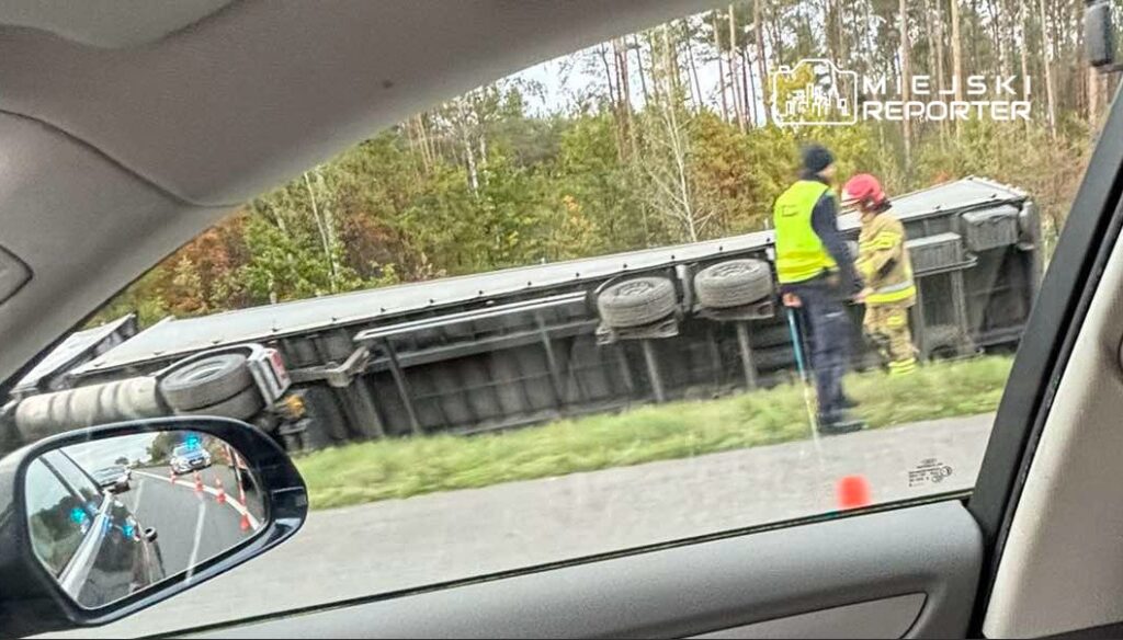 Na drodze leży przewrócony pojazd ciężarowy, a obok dwóch ratowników w odblaskowych kamizelkach ocenia sytuację.