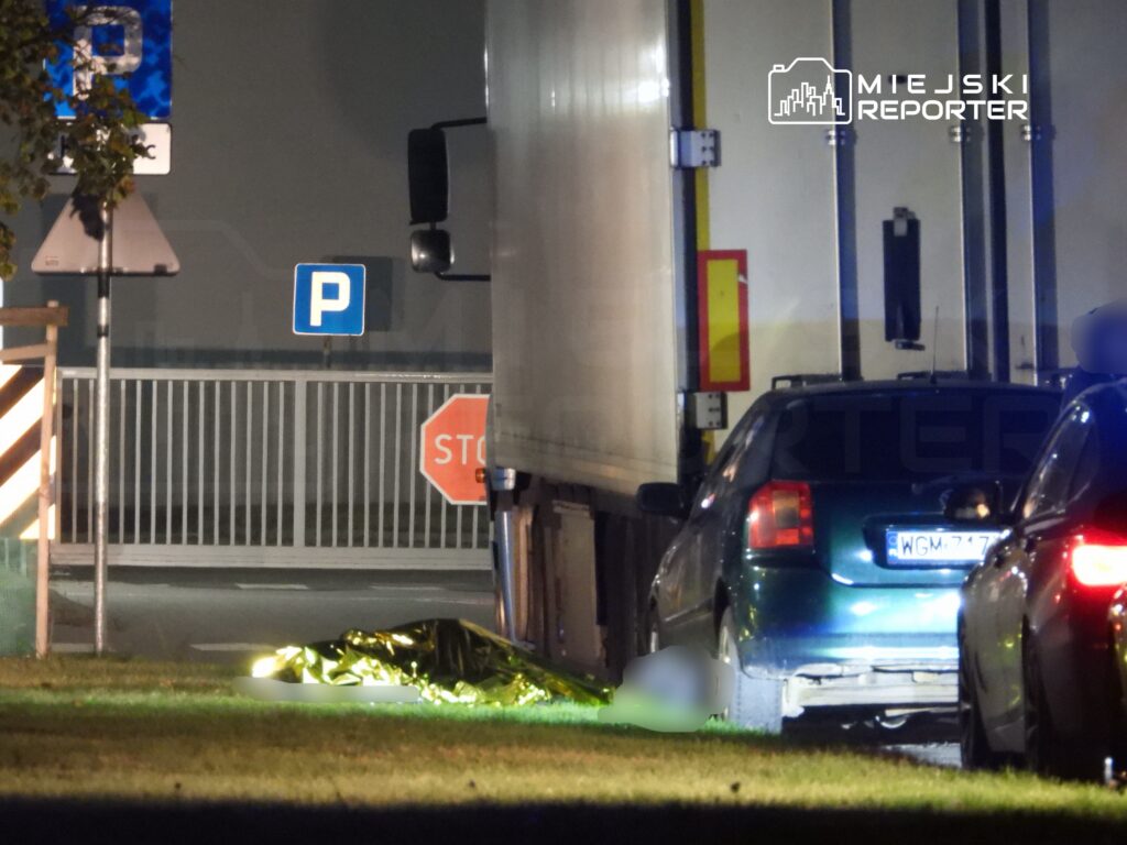 Złoty koc leży na trawie obok zaparkowanego samochodu, w tle widoczne znaki drogowe i ciężarówka.