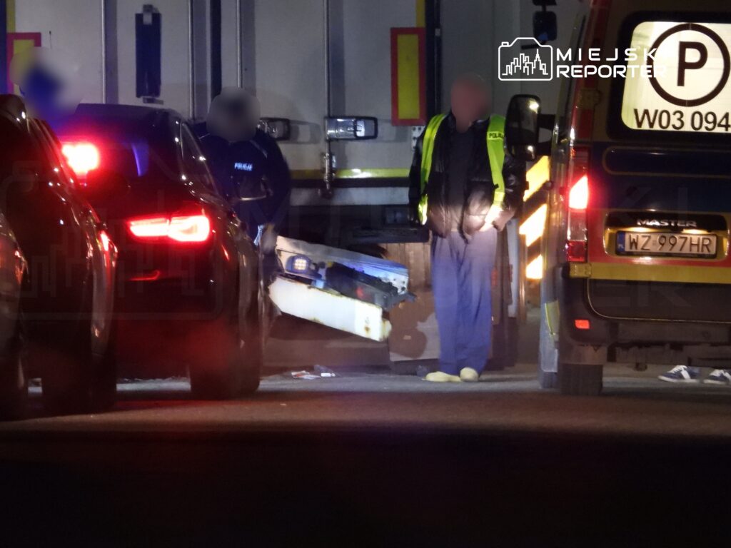 Policjant w odblaskowej kamizelce prowadzi czynności na drodze, obok stoi uszkodzony pojazd.