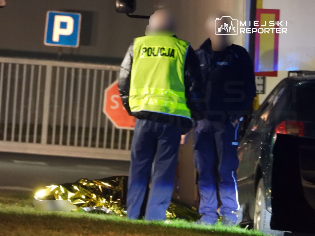 Policjanci w odblaskowych kamizelkach rozmawiają obok przykrytego folią ciała na parkingu.