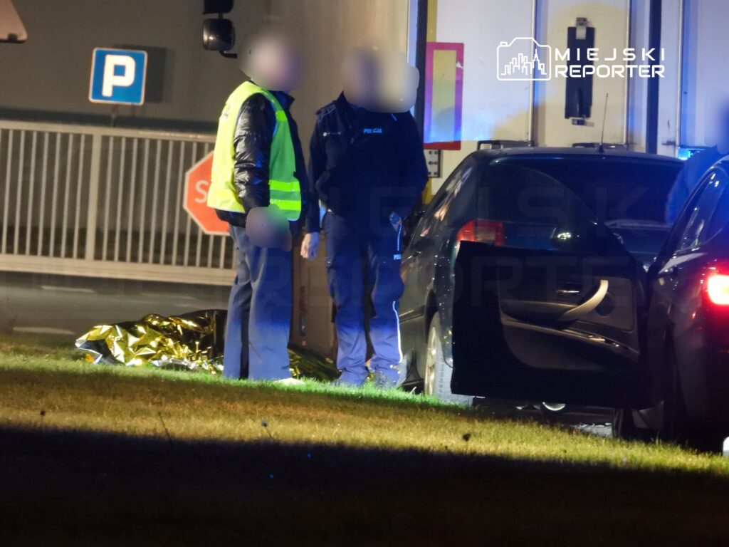 Funkcjonariusze policji rozmawiają obok przykrytej złotą folią postaci na trawie, w tle stoi czarny samochód.