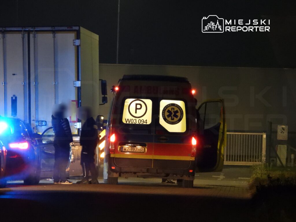Ambulans zaparkowany obok ciężarówki, a dwie osoby rozmawiają obok otwartych drzwi pojazdu. Nocna sceneria z niebieskimi światłami.