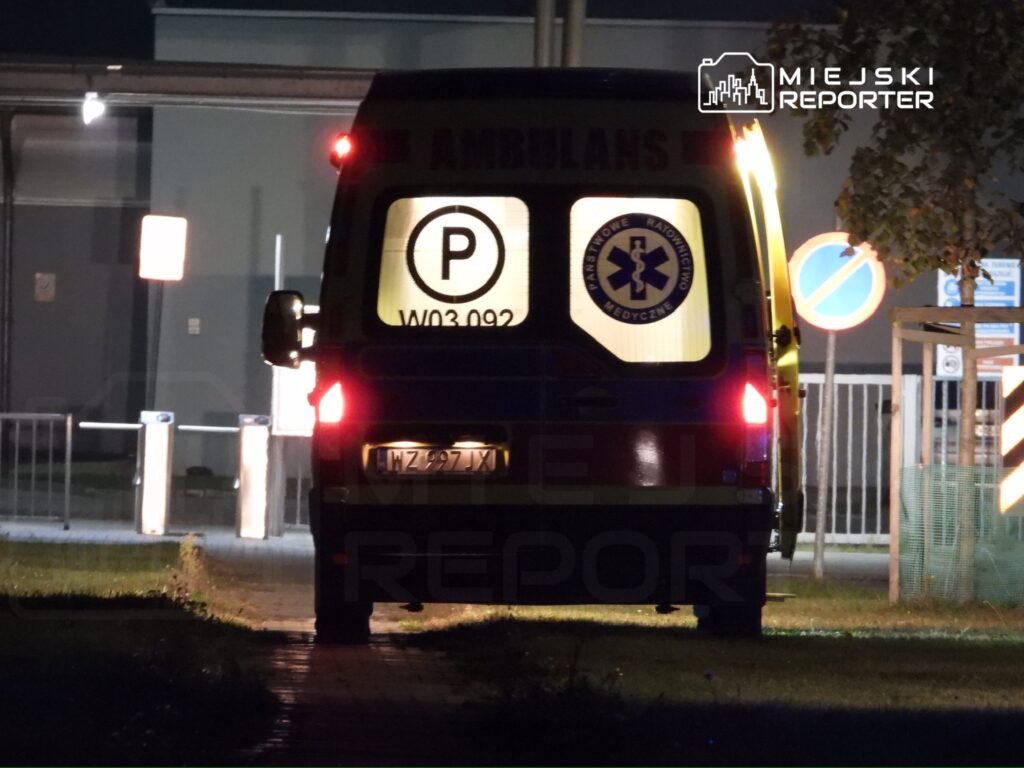 Karetka pogotowia z włączonymi światłami parkowania stoi na chodniku nocą, w tle widoczne znaki drogowe.