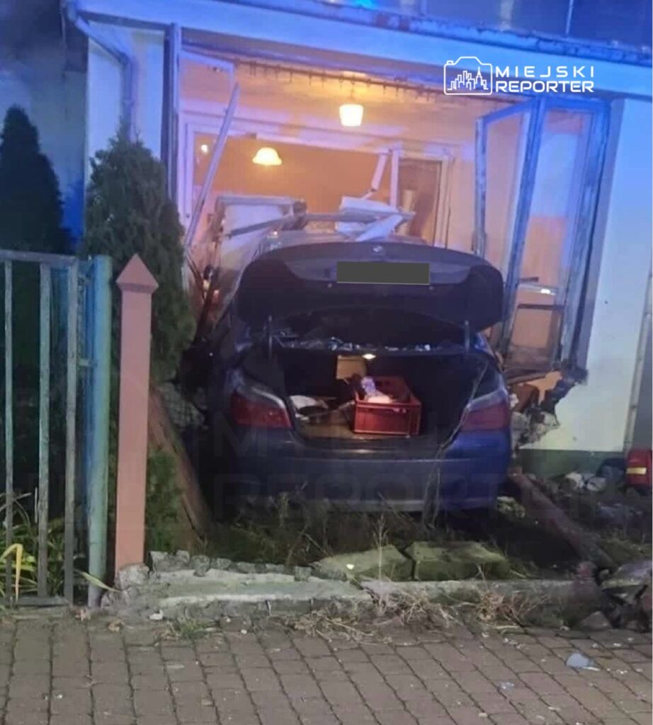 Samochód osobowy wjeżdża do budynku mieszkalnego, uszkadzając ścianę i okna. W tle widać zniszczenia wnętrza.