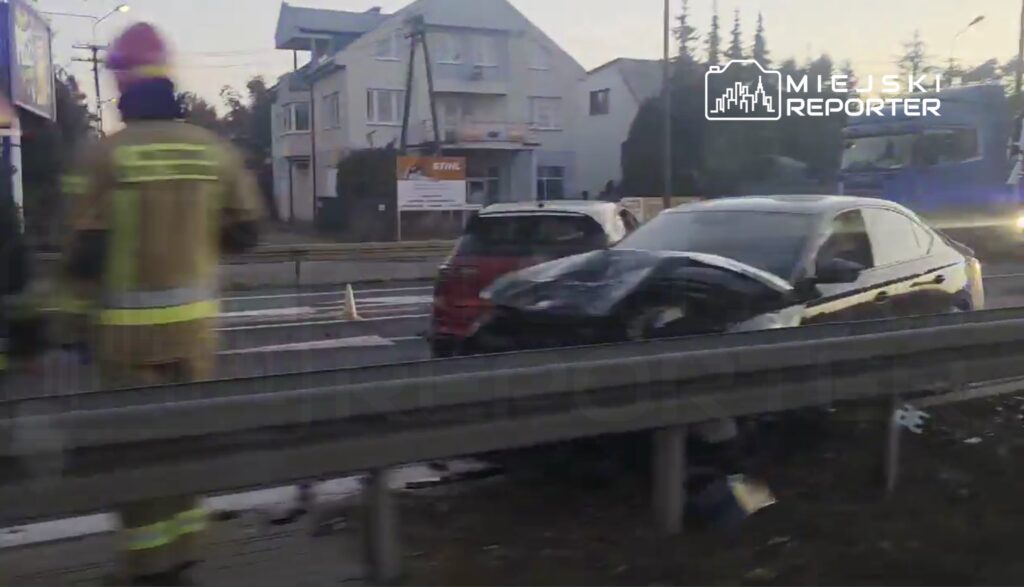 Strażak w mundurze z kaskiem ocenia uszkodzony samochód osobowy na poboczu drogi, obok stoi drugi pojazd.