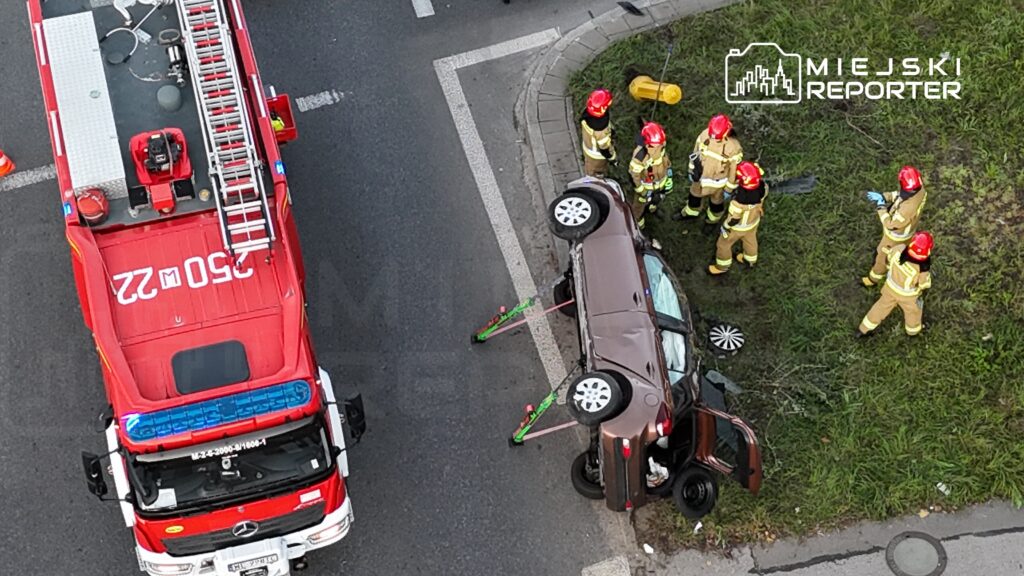 Ratownicy w mundurach pracują przy przewróconym samochodzie osobowym na skrzyżowaniu, obok wozu strażackiego.