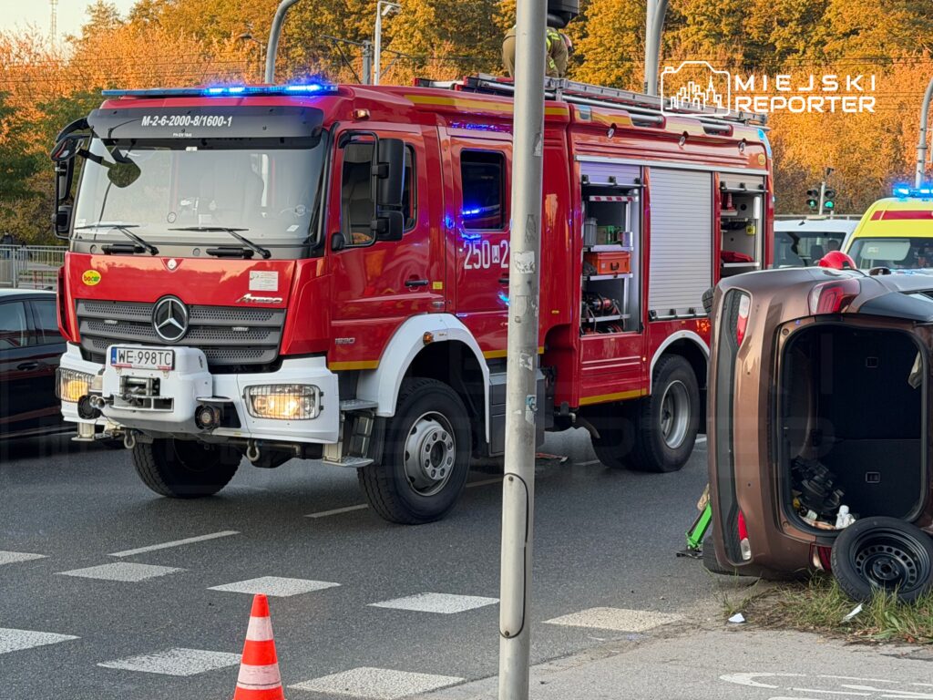 Czerwony wóz strażacki z włączonymi sygnalizatorami na skrzyżowaniu, obok przewrócone auto osobowe.