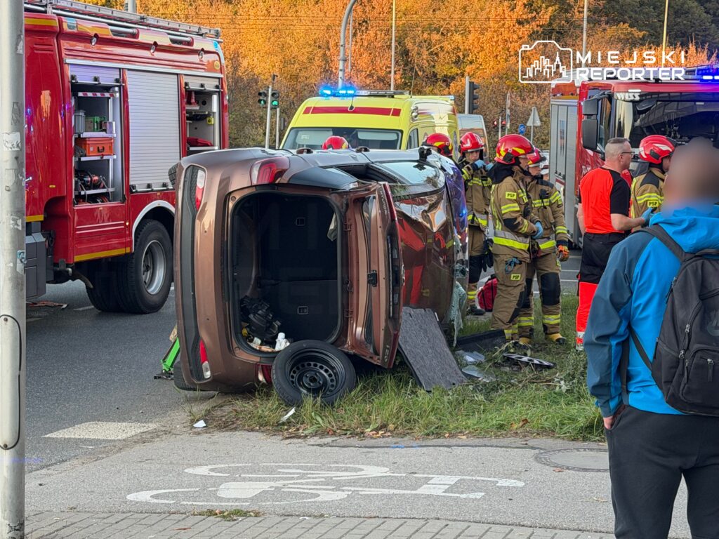 Na drodze leży przewrócone auto, obok grupka strażaków w mundurach oraz ratowników medycznych, w tle pojazdy ratunkowe.