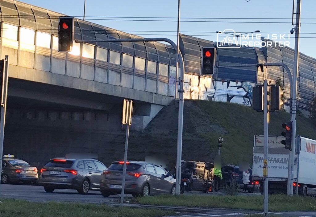 Na skrzyżowaniu stoją samochody, a w tle widać przewrócony pojazd i pracowników służb drogowych.