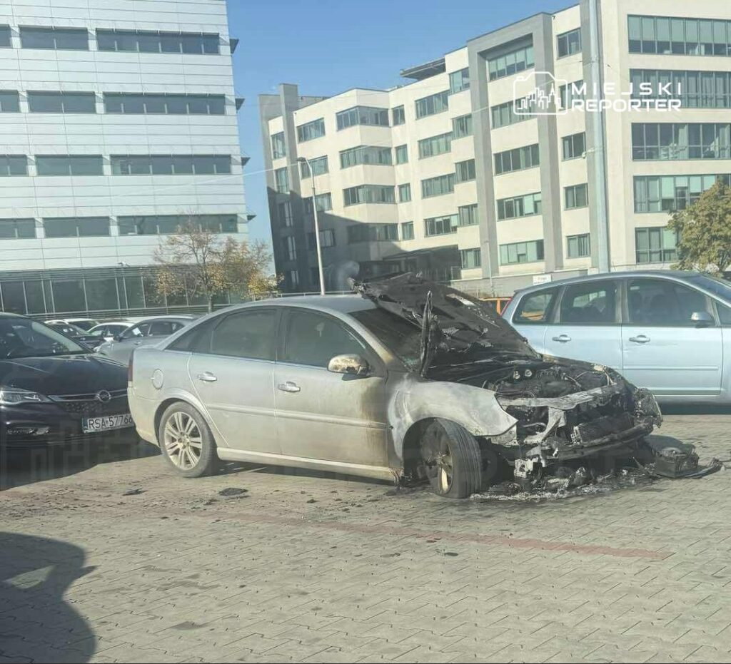 Zniszczony samochód z otwartą maską stoi na parkingu przed nowoczesnym biurowcem.