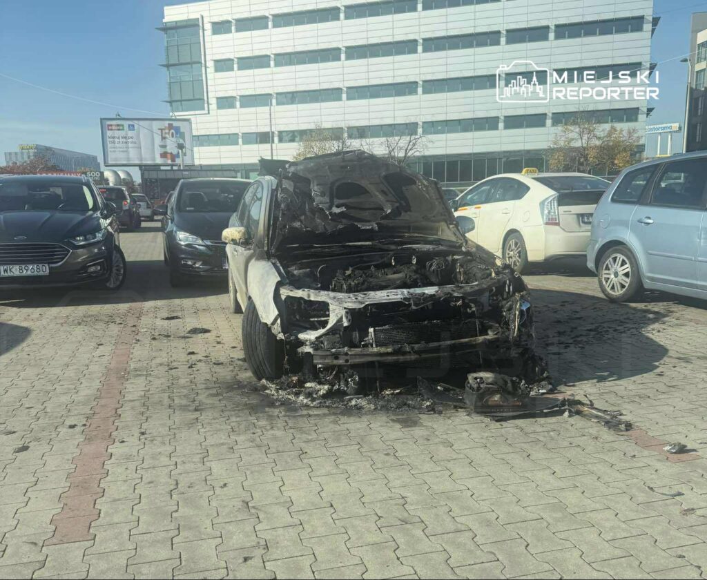 Spalony samochód osobowy stoi na parkingu, otoczony innymi pojazdami i nowoczesnym biurowcem w tle.