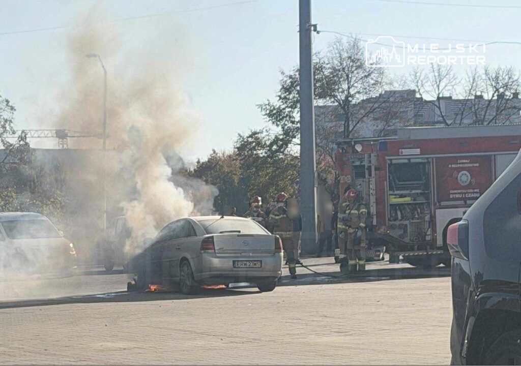 Samochód wydobywa dym z silnika, podczas gdy strażacy gaszą pożar w pobliżu.