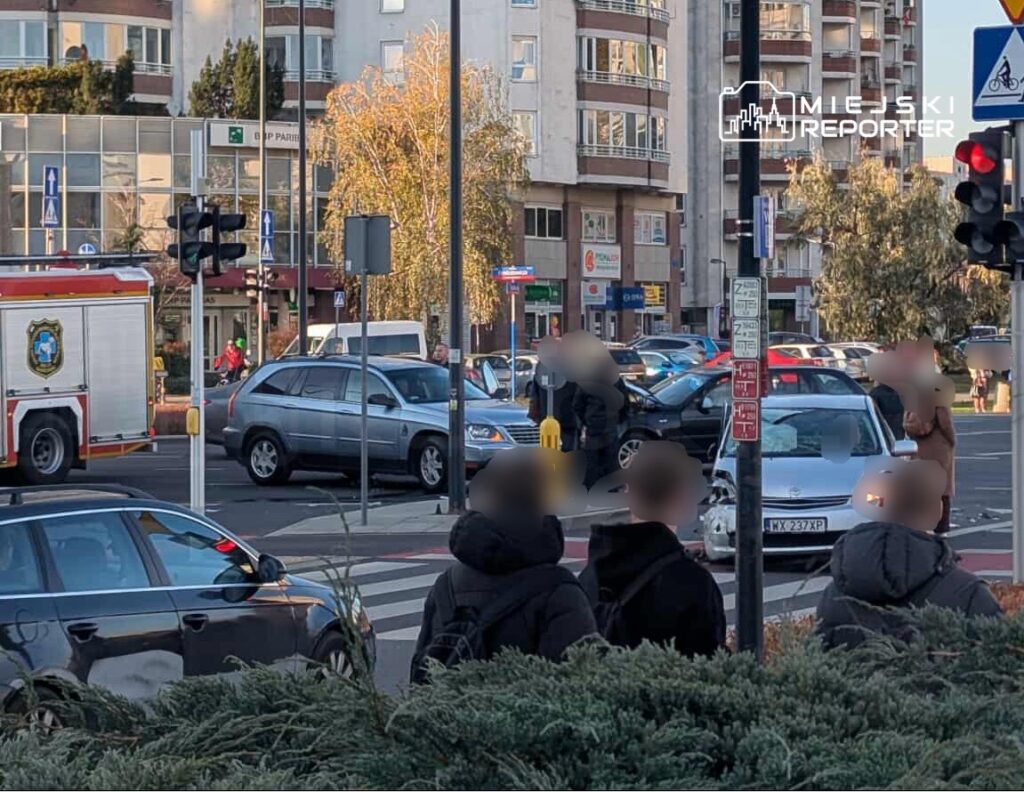 Wielu przechodniów obserwuje zderzenie dwóch samochodów na skrzyżowaniu, obok stacji straży pożarnej.