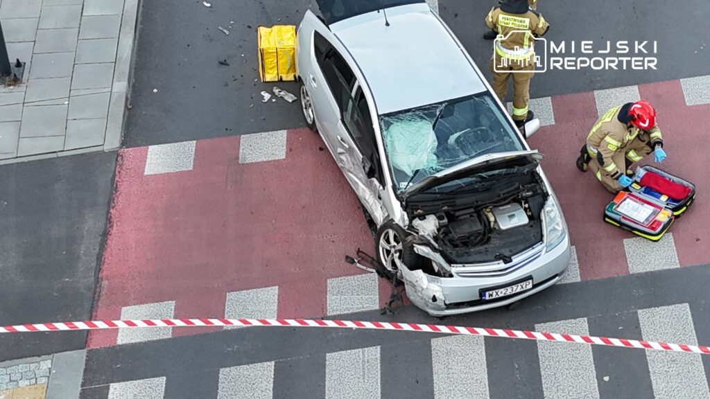 Uszkodzony samochód osobowy stoi na przejściu dla pieszych, obok interweniują strażacy z torbą medyczną.