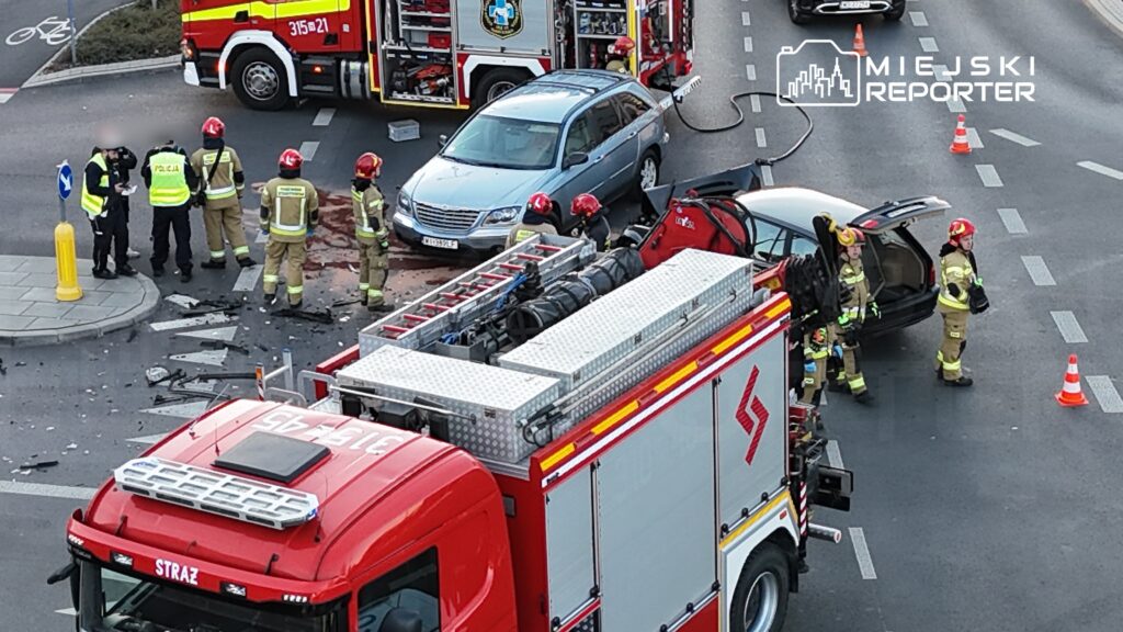 Grupa strażaków i policjantów pracuje na miejscu wypadku drogowego, obok pojazdu osobowego i wozu strażackiego.