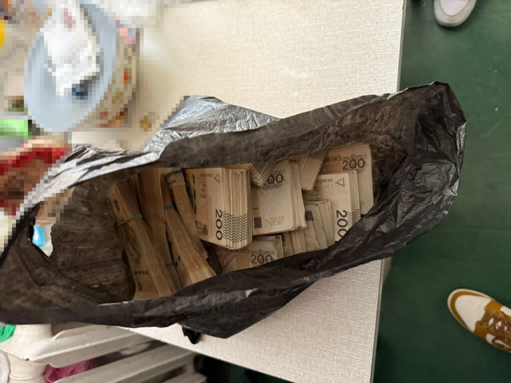 Czarna torba z zawartością banknotów o nominale 200 zł leży na stole w pomieszczeniu.