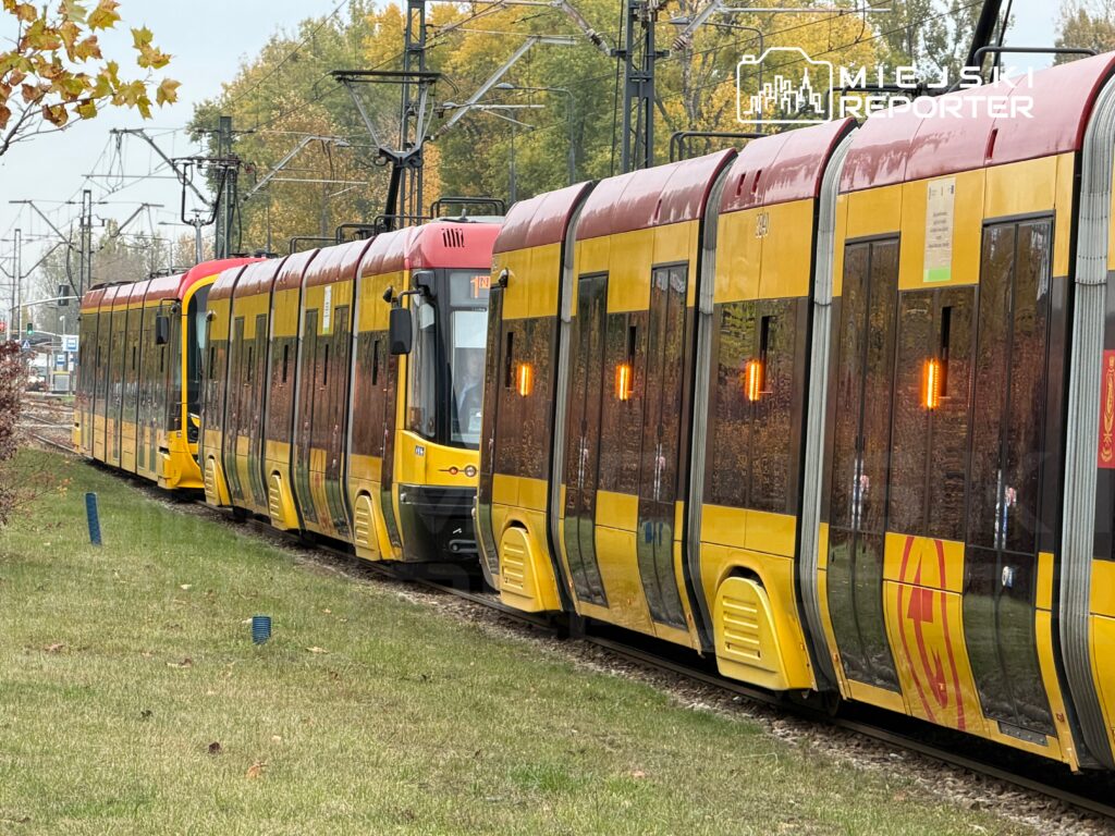 Tramwaj w kolorze żółtym i czerwonym zatrzymany na torach w otoczeniu zieleni i drzew, z widocznymi sygnalizatorami.