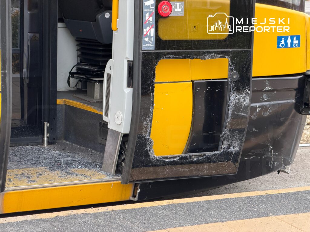Skrzynka z drzwiami tramwaju z uszkodzoną szybą i odłamkami szkła na podłodze przy przystanku.