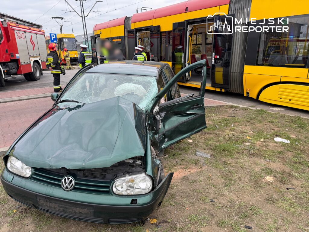 Zniszczony samochód osobowy stojący przy przystanku tramwajowym, obok strażacy i ratownicy medyczni.