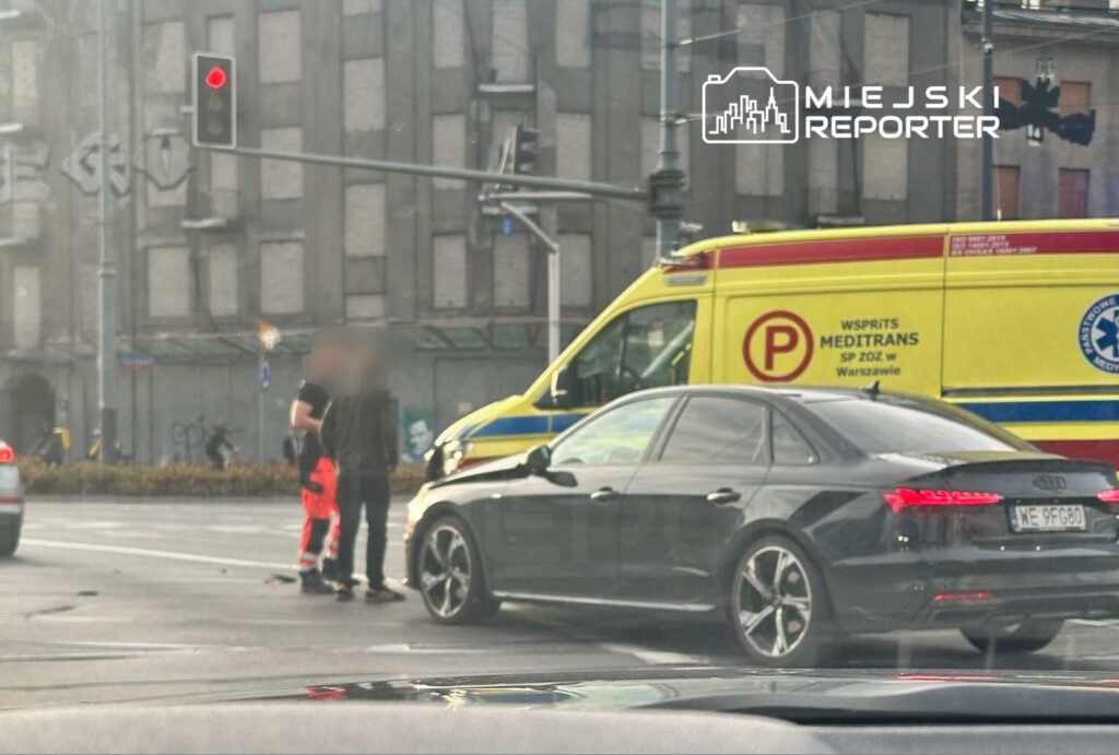 Mężczyzna w odzieży roboczej rozmawia z kierowcą czarnego samochodu osobowego na skrzyżowaniu, obok karetki pogotowia.
