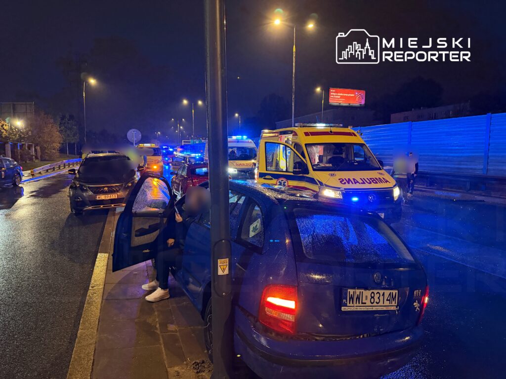 Na drodze w nocy zatrzymane są pojazdy, a przy nich ambulans z włączonymi sygnałami świetlnymi oraz osoby rozmawiające.