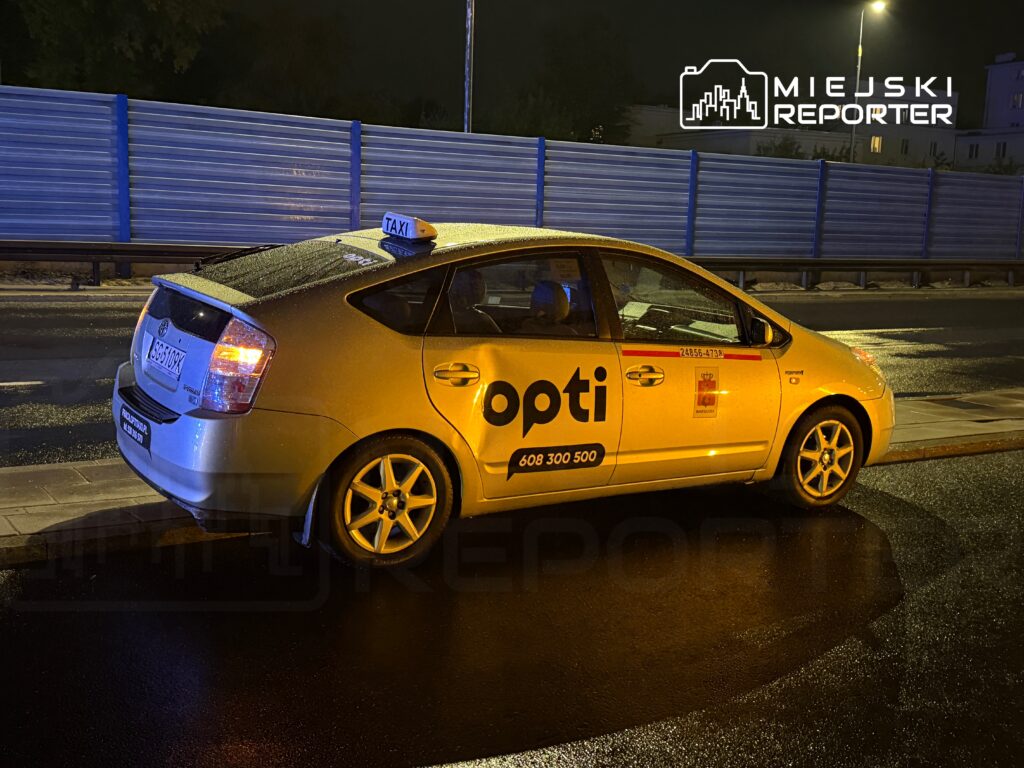 Żółte taxi marki Toyota Prius zatrzymane na mokrej nawierzchni, z widocznym napisem "opti" na drzwiach.