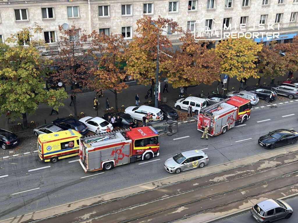 Strażacy oraz zespół ratownictwa medycznego pracują przy wozach strażackich na ruchliwej ulicy otoczonej drzewami.