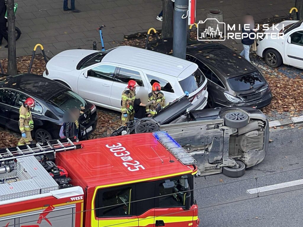 Strażacy w kaskach udzielają pomocy poszkodowanym przy przewróconym samochodzie na ulicy, obok wozu strażackiego.