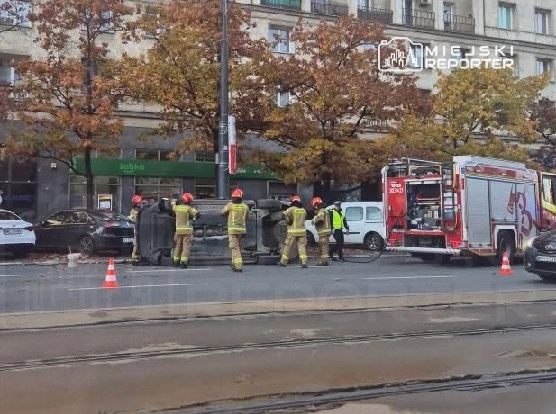 Grupa strażaków w hełmach próbuje postawić na koła przewrócony pojazd na ulicy, obok parkujących samochodów.