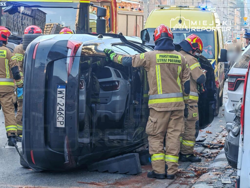 Strażacy w mundurach roboczych pomagają przy przewróconym na dach samochodzie osobowym na miejskiej ulicy.