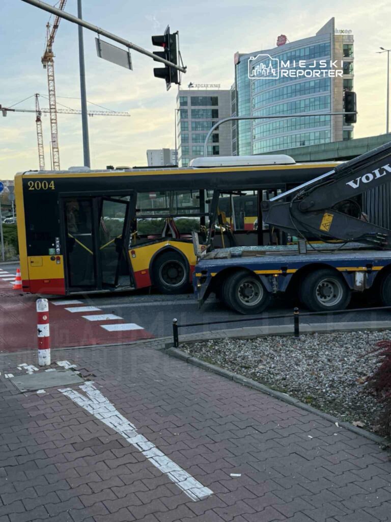 Autobus w żółto-czerwonych barwach stoi na skrzyżowaniu obok ciężarówki, w tle nowoczesne budynki biurowe.