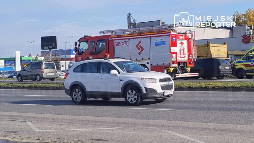 Biały samochód osobowy porusza się po drodze obok wozu strażackiego oraz karetki w miejskim otoczeniu.