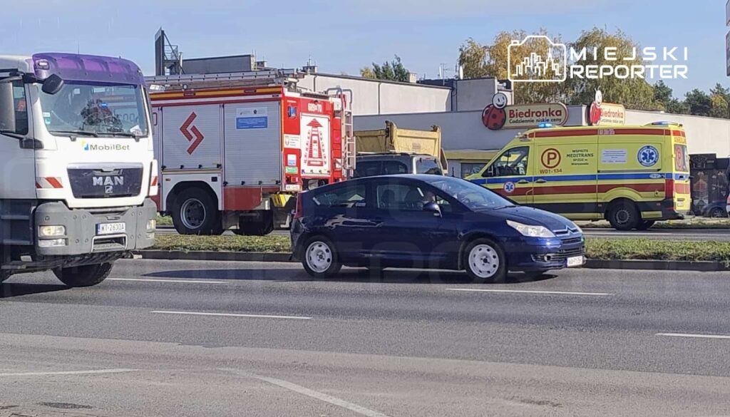 Czarny samochód osobowy porusza się obok wozu strażackiego i karetki na ruchliwej ulicy. W tle widoczny sklep.