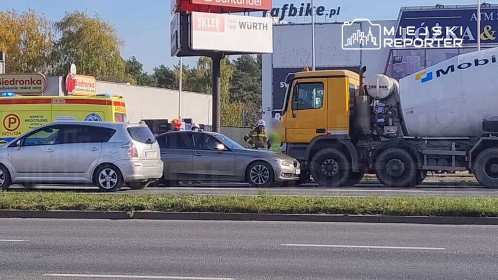 Wydarzenie drogowe z udziałem samochodu osobowego i ciężarówki, obok stacji benzynowej oraz ambulansu.