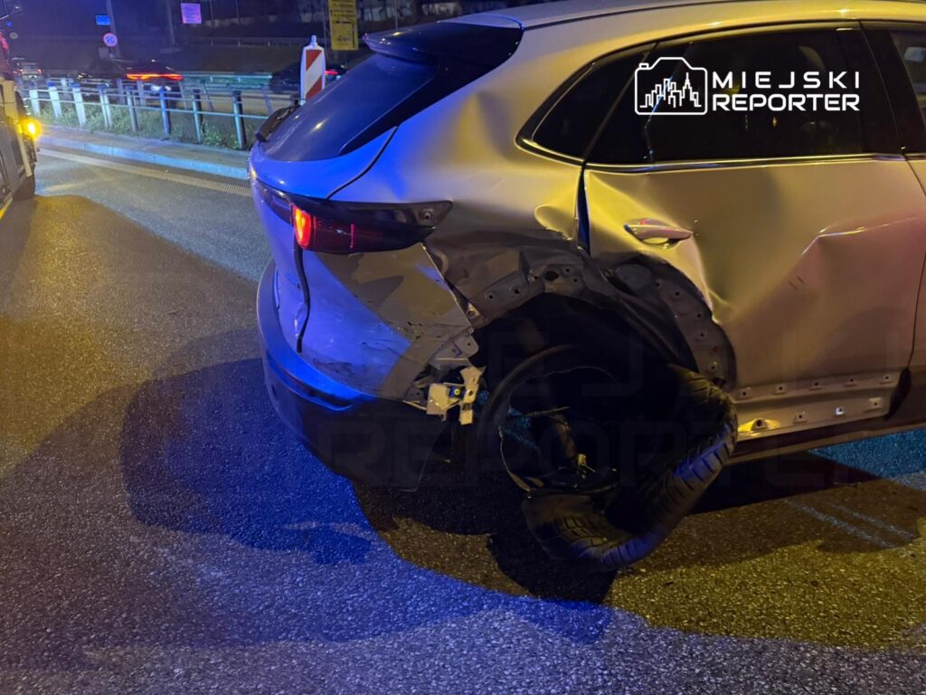 Uszkodzony samochód osobowy z oderwaną oponą stoi na poboczu drogi, obok widać inne pojazdy w ruchu.