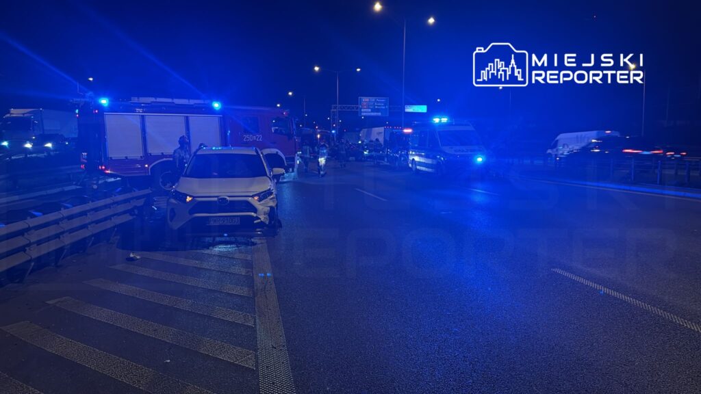 Na drodze nocą, białe auto z uszkodzeniem zderzaka, wokół pojazdy służb ratunkowych z włączonymi sygnalizacjami świetlnymi.