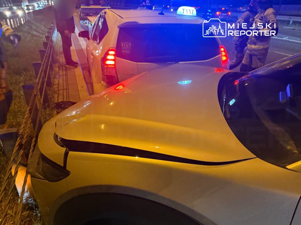 Dwa uszkodzone pojazdy na poboczu drogi, obok stoi mężczyzna w ciemnym ubraniu, a w tle widać inne samochody.