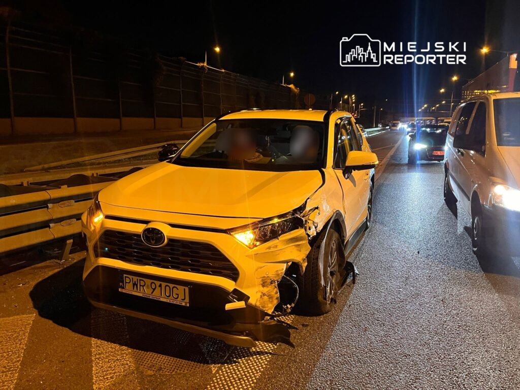 Uszkodzony biały samochód osobowy stoi na drodze nocą, obok barierki ochronnej, z widocznymi uszkodzeniami bocznymi.