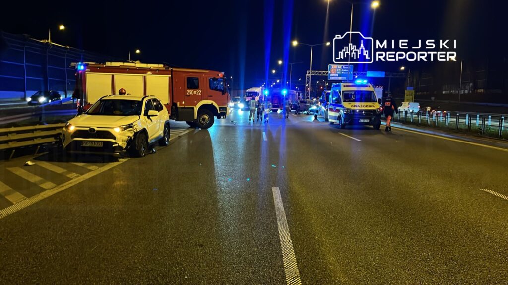 Na drodze nocą zderzenie samochodu osobowego z barierką; w tle widać pojazdy służb ratunkowych i policyjnych.