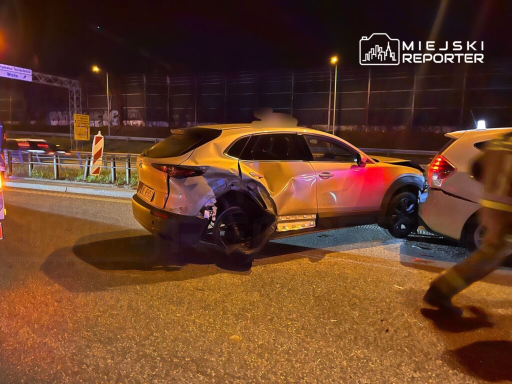 Uszkodzony samochód osobowy stoi na drodze, obok niego interweniujący strażacy w nocy.