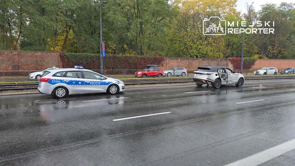 Policjant w radiowozie monitoruje sytuację na mokrej drodze, obok uszkodzony samochód z otwartymi drzwiami.