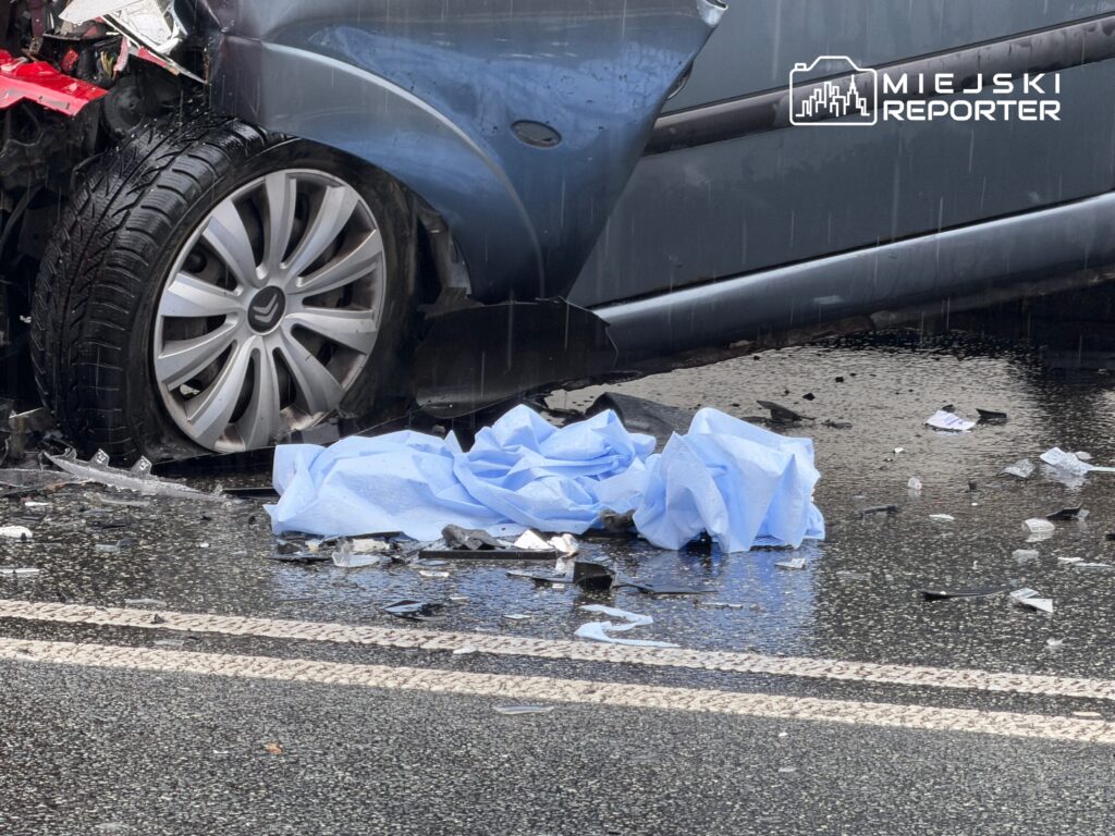 Zniszczony samochód osobowy leży na jezdni, obok rozrzucone fragmenty szkła i niebieska tkanina.