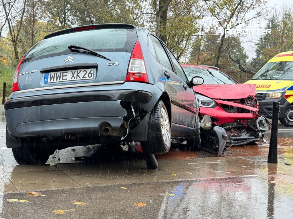 Dwa uszkodzone samochody osobowe, niebieski Citroen C3 i czerwony pojazd, zaparkowane na mokrej nawierzchni. W tle widoczna karetka pogotowia.