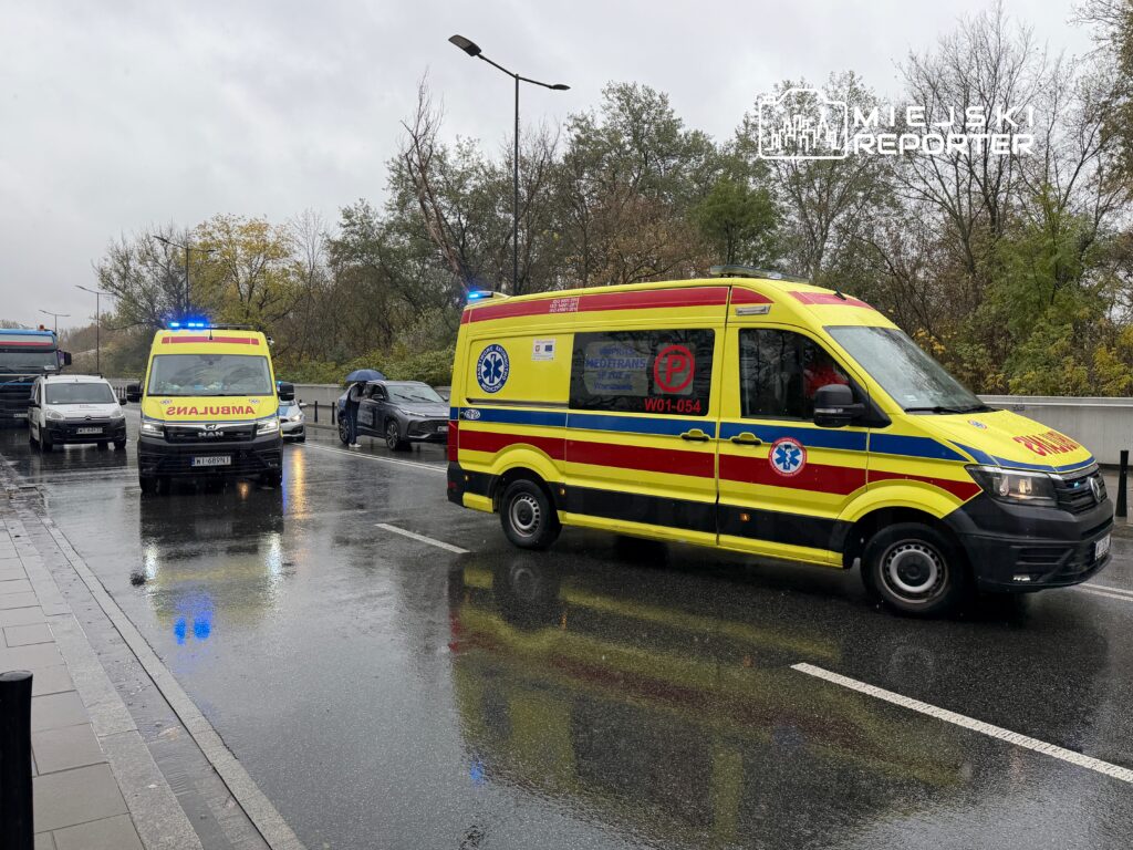 Ambulans z włączonymi sygnałami świetlnymi zatrzymany na deszczowej ulicy, obok innych pojazdów.