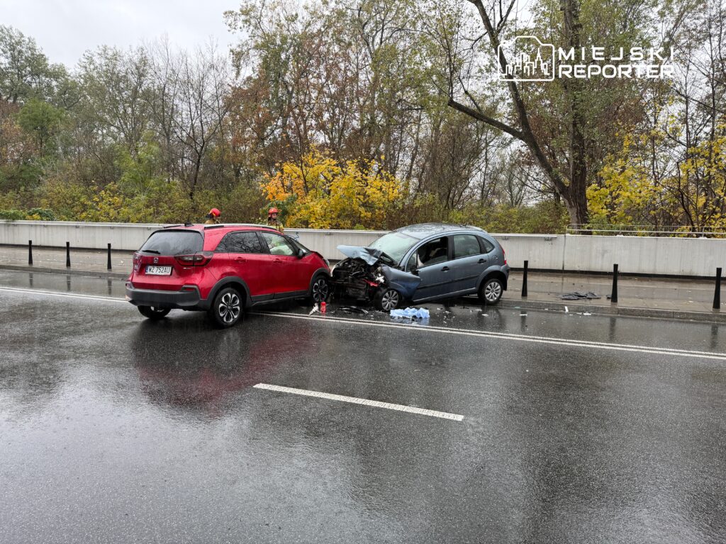 Czerwony SUV stoi obok rozbitego niebieskiego samochodu na deszczowej drodze, a w tle widać drzewa.