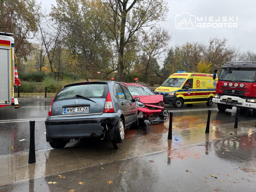 Dwa uszkodzone samochody osobowe na mokrej ulicy, obok pojazdy służb ratunkowych.