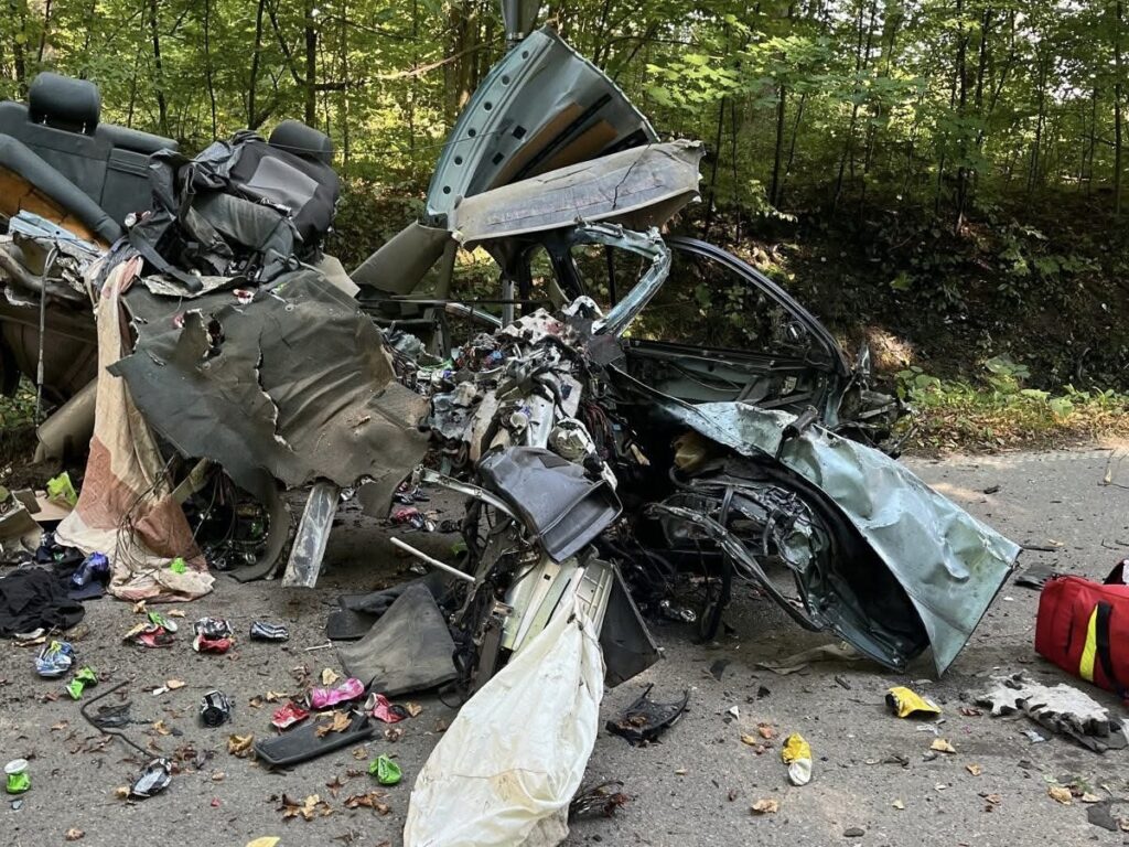 Zniszczony samochód osobowy leży na poboczu drogi, otoczony odpadkami i resztkami materiałów, w lesistym otoczeniu.