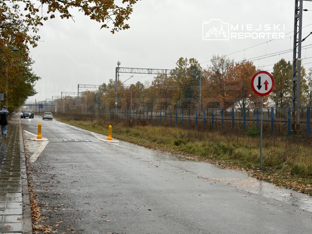 Kierowcy przejeżdżają obok znaku drogowym w deszczowy dzień na ulicy z torami kolejowymi w tle.